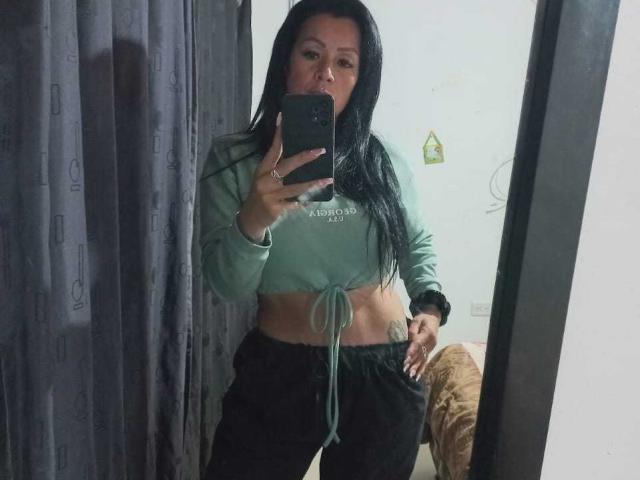 CelesteLaurent with long black hair, green crop top, tied at the front, black pants, holding phone in mirror selfie, showing a tattoo on her right hip.