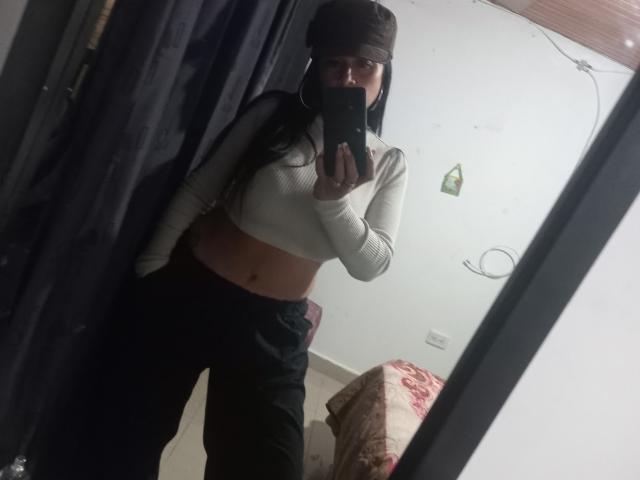 CelesteLaurent with long black hair, wearing a brown beanie, white long-sleeve crop top, and black pants, taking a mirror selfie, showing her midriff and belly button.