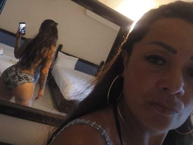 CelesteLaurent with long brown hair, hoop earrings, and a tattooed arm, wearing fishnet shorts and a tank top, taking a selfie in a mirror, showing her butt.