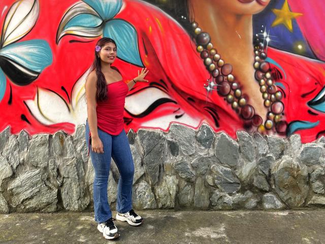 AlyssaGreen stands in front of vibrant graffiti. She's a Latina woman with long brown hair, wearing a red strapless top, blue jeans, and white sneakers. Her right hand is raised in a wave. The graffiti features a large red face with floral elements, a close-up of a woman's face with a star and beads, and a rocky texture at the bottom. She has a purple flower in her hair. The colors are bold and the textures contrast.