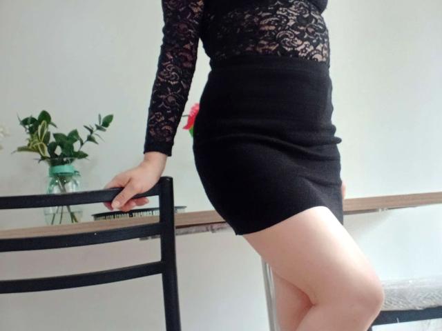 ScarletMilfX with lace top, black skirt, and bare legs, leaning on chair, holding vase with green plants, showing her ass and thighs.