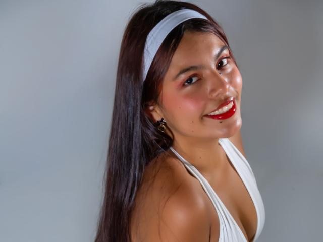 AlyssaGreen with long brown hair, white headband, red lipstick, white halter top, smiling, showing her tits, small earrings, light skin, glossy red lips, looking over her shoulder.