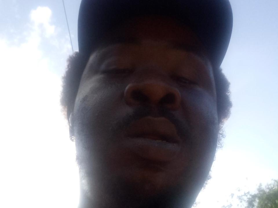 Prikev69's dark brown face with a curly mustache, wearing a black cap, in a low-angle shot, showing their nose and slightly parted lips. Sunlight highlights their skin.