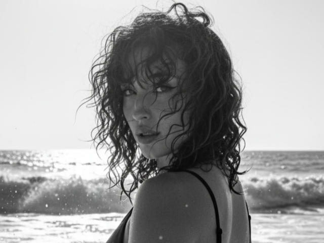 Black-and-white photo of NickiCandy with wet, curly hair, looking back over her shoulder, wearing a black spaghetti-strap top, showing her small tits and a slightly open mouth.