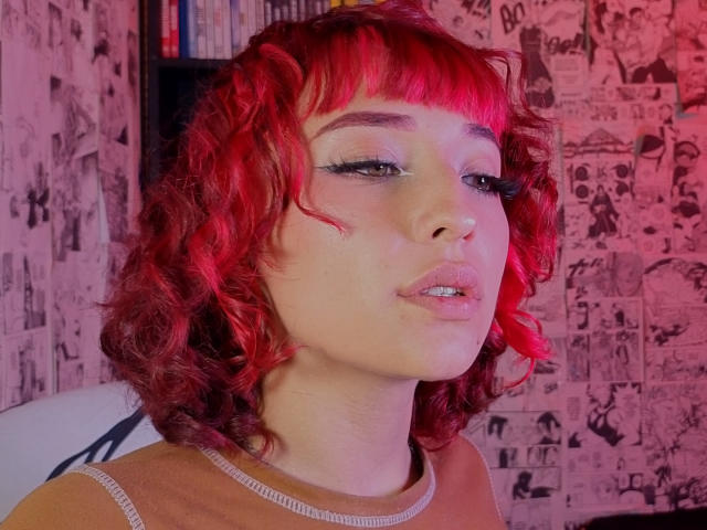 NickiCandy has red curly hair, eyeliner, and pink lipstick. She's wearing a brown sweater, looking slightly to the side with parted lips. Her skin is fair.