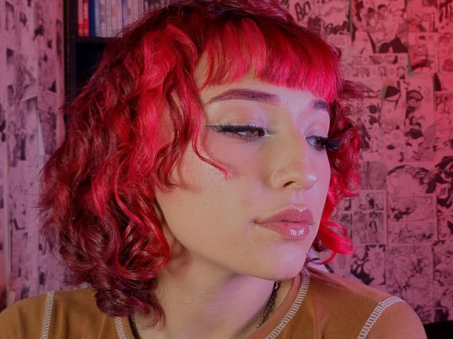 NickiCandy with bright red curly hair, pale skin, and shiny pink lips, wearing a brown top, has red-lit makeup and a nose ring, with eyelashes slightly open.