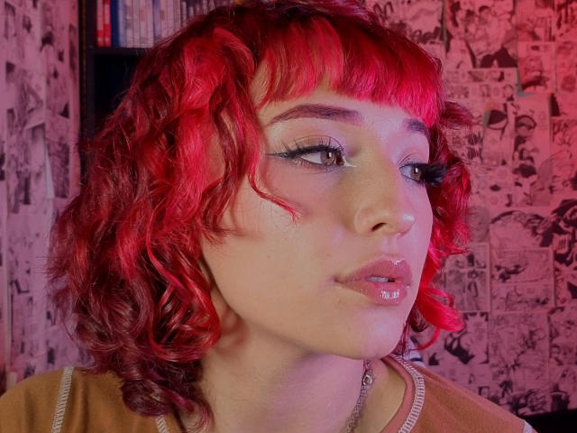 NickiCandy with red curly hair, pink glossy lips, and white eyeliner, looking to the side, has a shiny nose and a necklace, in a red-lit photo.