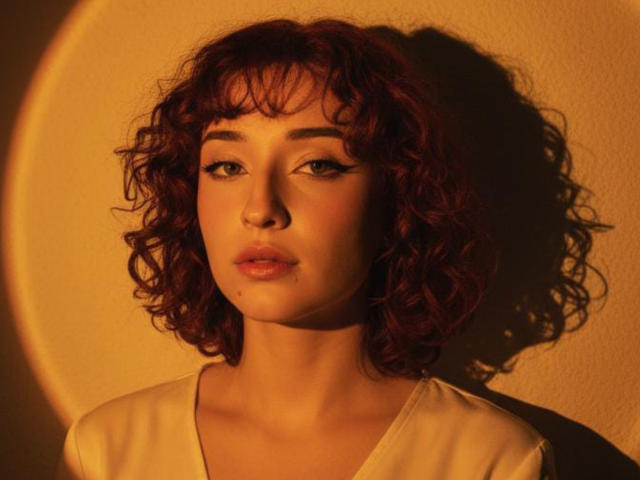 NickiCandy has curly red hair, light skin, and is wearing a white shirt. She's looking slightly to the side with a serious expression, bathed in warm, golden light.