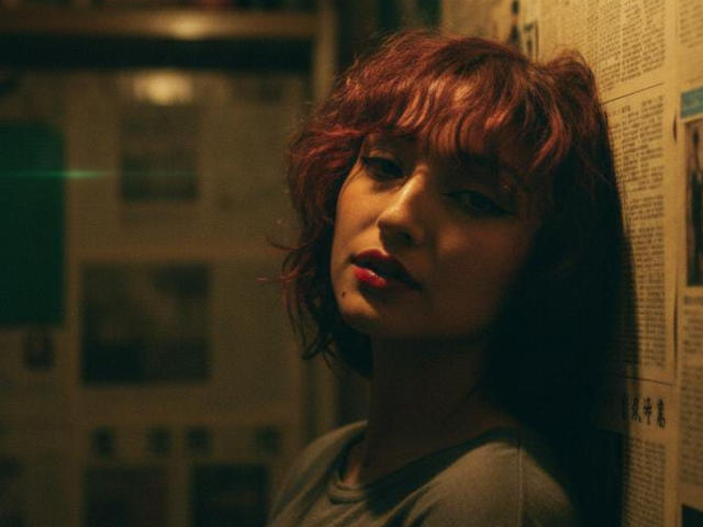 NickiCandy with messy red hair, dark eyeliner, and pink lipstick, leaning against a wall, wearing a gray shirt, looking tired and slightly annoyed, in a dimly lit room.