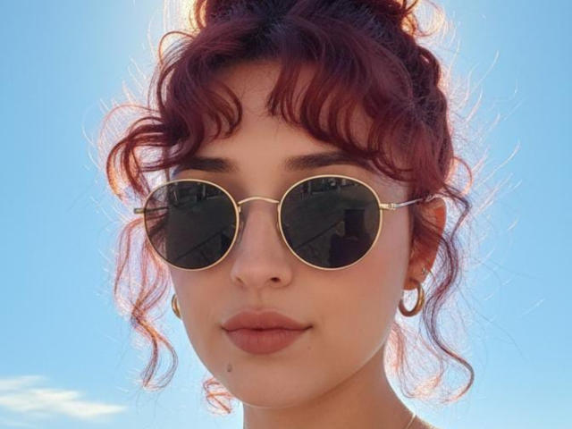 NickiCandy has red curly hair, round gold-rimmed sunglasses, and gold hoop earrings. Her fair skin and pinkish lips are visible against a bright blue sky.