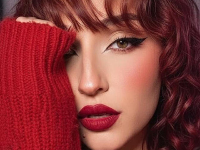 NickiCandy has red curly hair, red lipstick, and is winking with her right eye, covered by a red knitted finger, showcasing black eyeliner and a cute lip piercing.