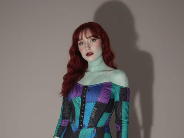 NickiCandy with pale blue skin, red wavy hair, dark lipstick, wearing an off-shoulder, colorful, checkered dress, buttons down, shadow on the wall.