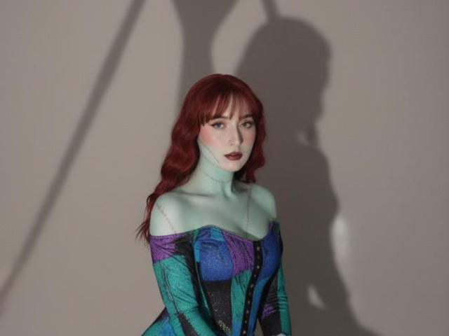 NickiCandy has pale green skin, red wavy hair, and wears a colorful off-shoulder dress with purple and blue patterns. Her shadow is on the wall.