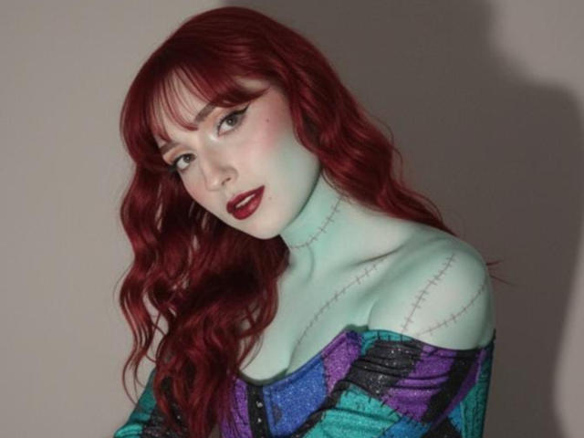 NickiCandy has pale green skin, red wavy hair, dark eyeliner, and purple lipstick. She's wearing a colorful off-shoulder top with visible stitches, showing her tits.