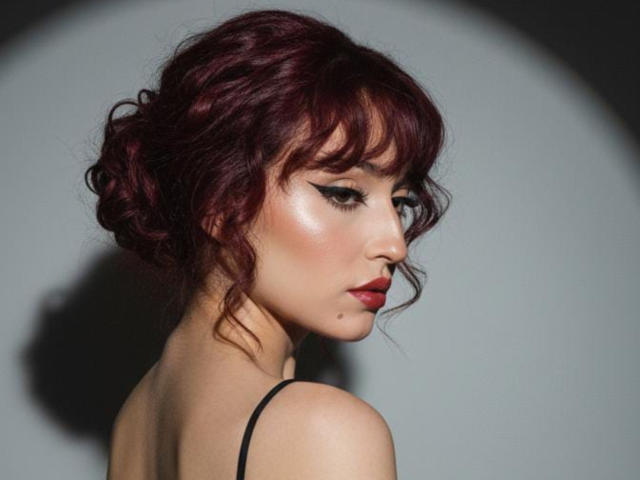 NickiCandy with red curly hair, black eyeliner, red lipstick, and a mole on her chin, looking to the side with a shadow on her left shoulder.
