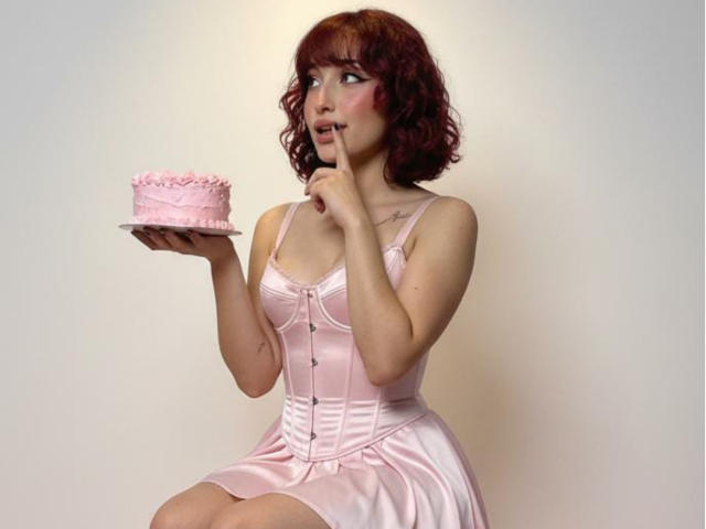 NickiCandy with red curly hair, wearing a pink satin dress, holding a pink cake, finger to her lips, looking surprised. Her tits are visible under the dress.
