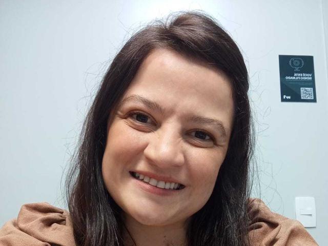 Close-up of LuxEsa with a slight tilt to the right. She has fair skin, dark brown, slightly messy hair, and a subtle smile. Her eyes are brown, and she has light makeup. She wears a brown, fuzzy-textured top. The background is a plain, light gray wall with a green sticker on the upper right showing "Infection Control" and a QR code. The image has a casual, slightly imperfect feel.