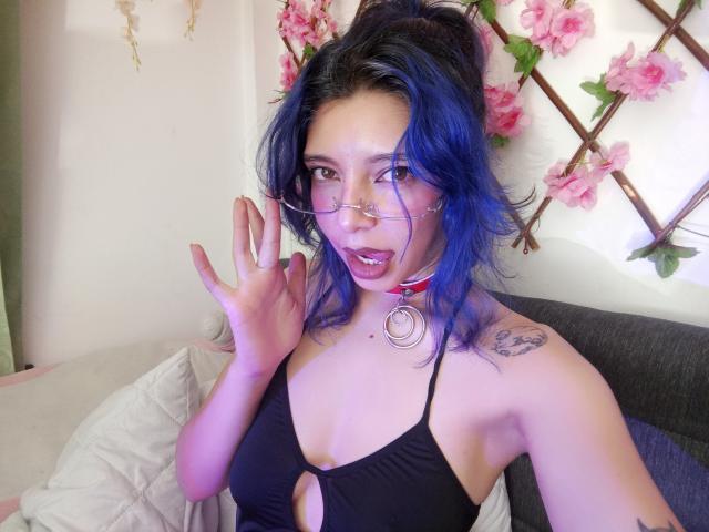DarckVelvet has blue-dyed, shoulder-length hair and a fair skin tone. She wears a black, cutout top revealing cleavage, and a red choker with a metal ring. Her right hand is raised, showing a silver ring on her finger. She has a nose ring and a tattoo on her right shoulder. Pink cherry blossom decorations are on the wall behind her. Her expression is slightly open-mouthed, looking at the camera. The image has a casual, modern aesthetic.