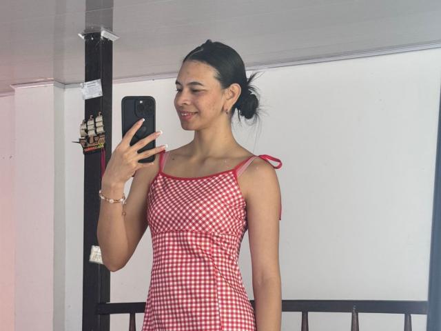 AmberGray69 with small tits, wearing a red and white checkered dress, holding a phone, smiling, black hair in a bun, standing in a room with a black pole.