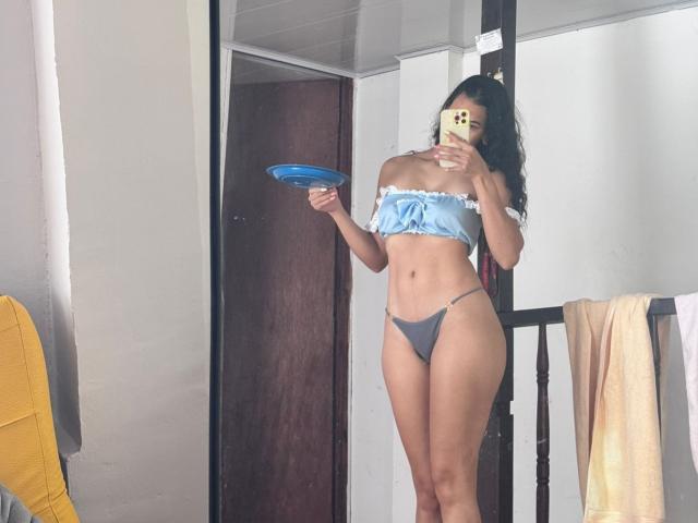 AmberGray69 with long black hair, wearing a blue off-shoulder top and gray thong, holding a blue plate, mirror selfie, showing her tits and ass, standing in a room.