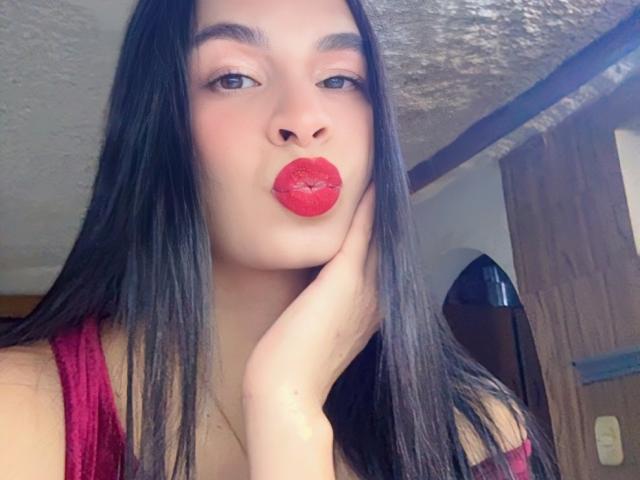 AmberGray69, con cabello negro largo, lápiz labial rojo y un vestido rojo, besando la cámara con una mano en la mejilla, mostrando senos grandes y un rostro lindo, en una habitación luminosa..
