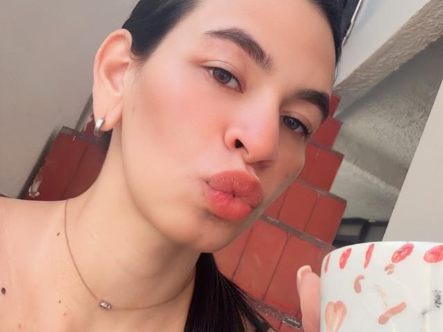 AmberGray69, con labios fruncidos, ojos marrones y cabello oscuro, lleva un collar, sostiene una taza blanca con diseños rojos, tiene pequeños aretes y un ligero bronceado..