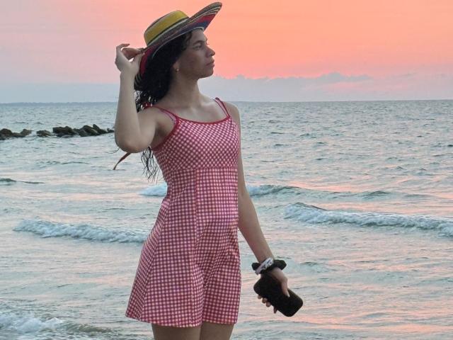 AmberGray69 con un vestido a cuadros rojo y blanco, sosteniendo una cámara, usando un sombrero de paja, de pie junto al océano, con cabello castaño ondulado, y un atardecer en el fondo..