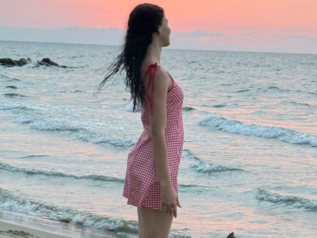 AmberGray69 in a red and white checkered dress, standing side profile at the beach, with wavy black hair, small tits, and a cute ass, looking at the sunset.
