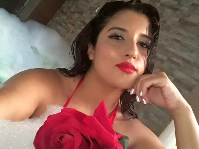 AlisRoberts with wet black hair, red lipstick, and a red rose halter, in a bubbly bathtub, resting her chin on her hand, looking at the camera.