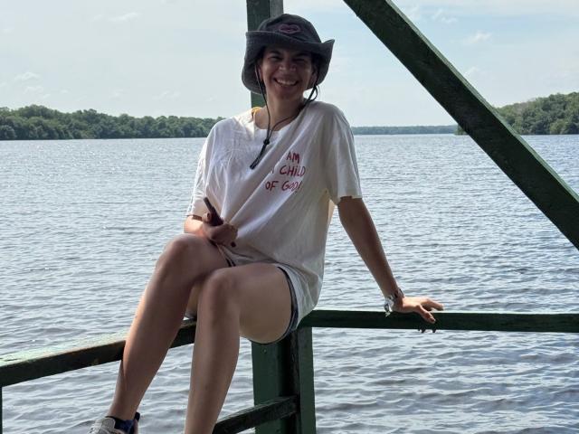 AmberGray69 está sentada en una barandilla verde, vestida con una camiseta blanca y pantalones cortos, un sombrero, sonriendo, con las piernas cruzadas, mostrando su trasero, cerca de un lago..