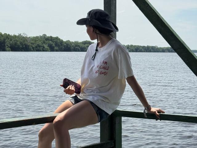 AmberGray69 sits on a green wooden railing, wearing a black hat, white shirt, and denim shorts, holding a phone, with a forested shoreline in the background.