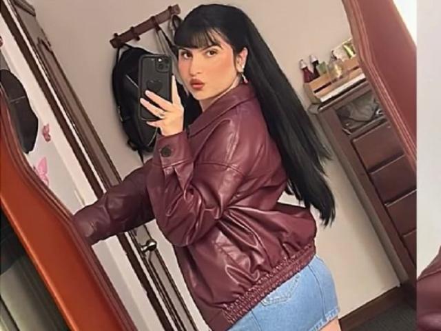 EmilySpaceI with long black hair, wearing a maroon leather jacket and blue denim shorts, taking a mirror selfie, showing her ass, holding a black phone, in a bedroom.