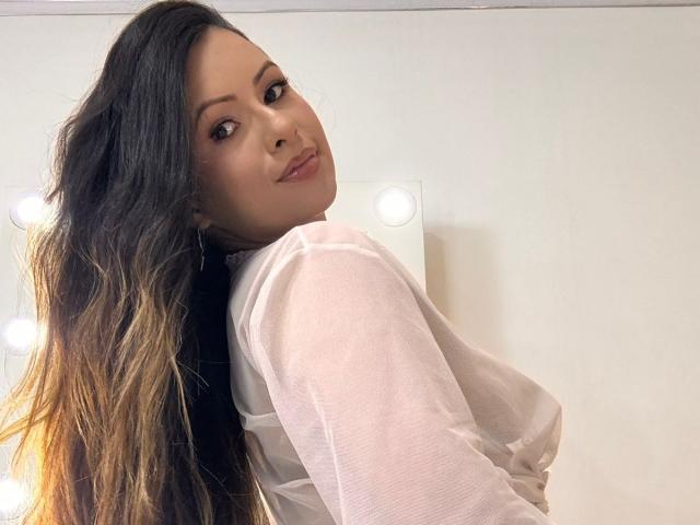 HannaRobertss with long, wavy brown hair, light skin, and subtle makeup, wearing a sheer white top, looking over her shoulder with a slight smile. Boobs visible through top.