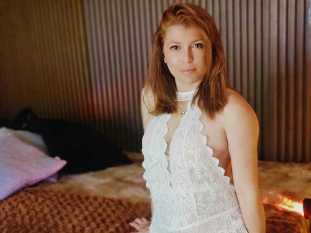MoonXVelvet, a light-skinned woman with shoulder-length brown hair, stands in a room. She wears a white, lace, halter-neck lingerie with scalloped edges and a deep V-cut. Her eyes are slightly downcast, and she has a neutral expression. The background features a bed with a brown, textured blanket and a pillow to the left. Vertical, ribbed wall panels are behind her. The lighting is warm, highlighting her skin and the lace's intricate pattern. She is centered in the frame, with her right hand resting on the bed.