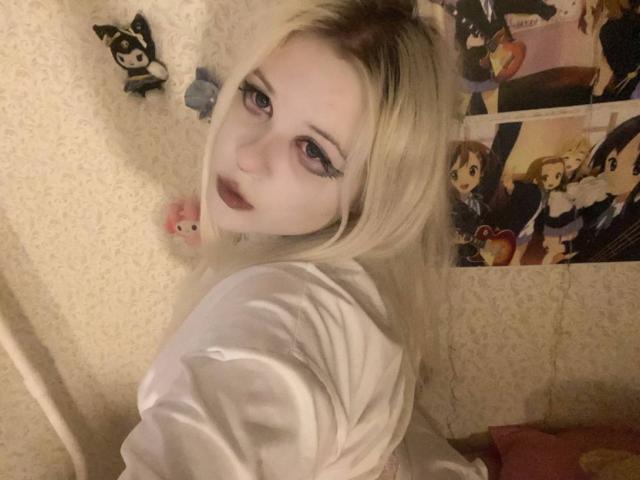 TinaEvanss with pale skin, blonde hair, dark lipstick, and heavy eyeliner, wearing a white shirt, looking over her shoulder, surrounded by anime figurines and posters.