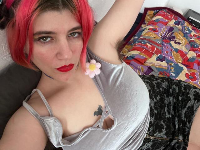 RozyReed-hot with pink hair, red lipstick, grey top, black lace shorts, flower in hair, arm raised, tattoos, looking at camera, sexy, visible cleavage, black ear gauges.