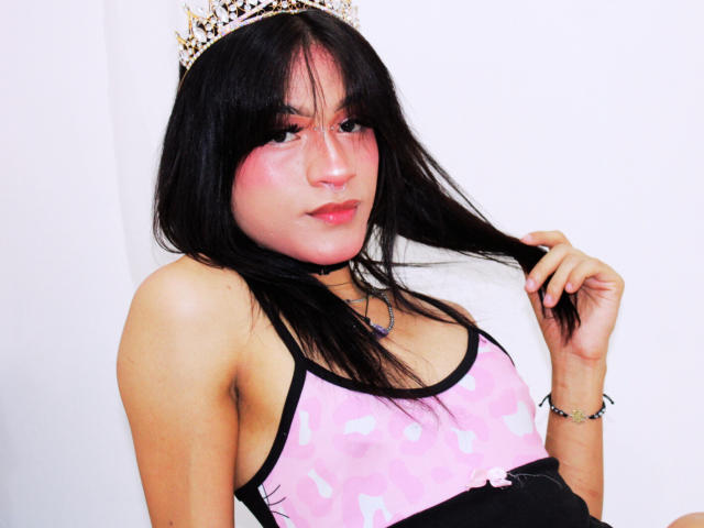 MoonlyVelvett has black hair, pink blush, and wears a pink camo top with black trim, a choker, and a sparkly crown, holding her hair with a cute smile.