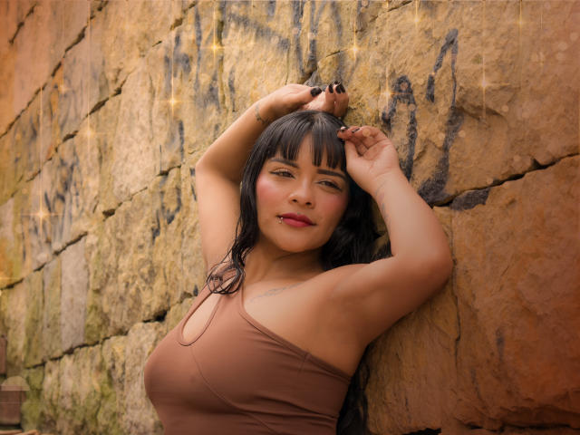 MiaBellucci with wet black hair, arms above head, brown tank top, showing tits, against a stone wall with graffiti, slightly flushed face, red lipstick, black nails, confident look.