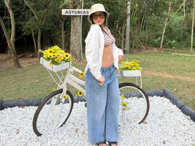 MiiaPalmer stands in a gravel-filled area with a white bicycle, flower pots filled with yellow flowers on either side of the bike. She wears a beige bucket hat, large sunglasses, a striped crop top, and loose blue jeans. Her midriff is exposed. The bicycle and flower pots are positioned behind her, with a sign reading "ASTURIAS" above. The image has a casual, outdoor, summer vibe. Her stance is relaxed, with hands near her waist. The background is a forested area.