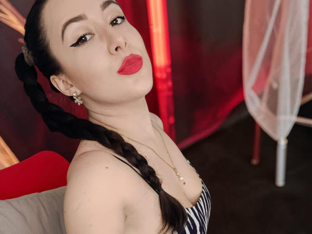 MabyLegrand has fair skin, dark braided hair, and is wearing bold red lipstick. She has black winged eyeliner, small star earrings, and a gold necklace with a small pendant. She's dressed in a black-and-white striped top, showing a bit of cleavage. She's seated with a red and gray chair in the background, slightly turned to the left. The lighting is warm, highlighting her facial features and red lips.