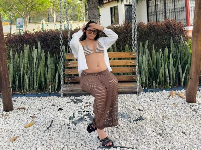 MiiaPalmer sits on a wooden swing in a pebble-covered area. She wears a white open shirt, striped bikini top, and brown fishnet pants. Her hands touch her head, and she wears black sandals. She has dark hair and wears sunglasses. The background includes green plants, a fence, and a building. The pebble ground contrasts with the wooden swing. Her relaxed pose and casual outfit create a laid-back vibe. The image uses natural light.