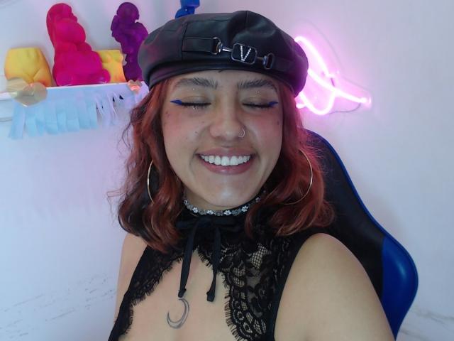 CuteH with red hair, wearing a black leather cap with glasses on it, black lace top, and large hoop earrings. She's smiling with eyes closed, showing a nose ring and blue eyeliner. She's seated in a blue chair, with a neon pink light behind her. The background has colorful sex toys on a shelf. Her face is slightly messy with white fluid on her chin.