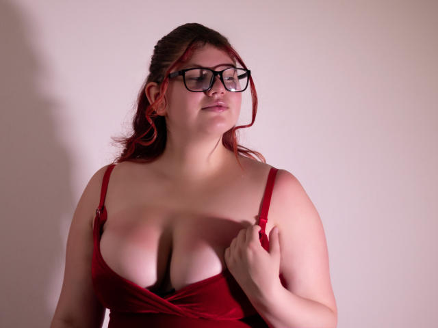 JohannaCharm, with red hair and glasses, holds her red dress, showing her big, juicy, bare-ass tits. She's fair-skinned, looking down, in a soft, pink-lit setting.