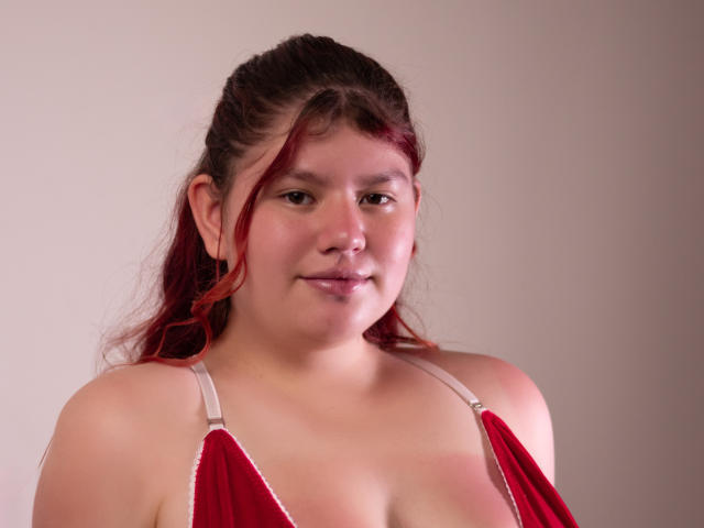 JohannaCharm with red velvet top, showing her tits, has a slightly chubby face, light skin, and red-streaked brown hair, smiling softly, against a plain background.