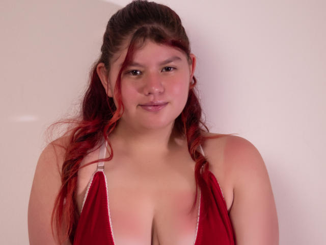 JohannaCharm, red-haired, plus-sized woman, wearing a red, deep-pleated, sleeveless top with white straps, showing her large, bare tits, smiling softly, against a plain white background.