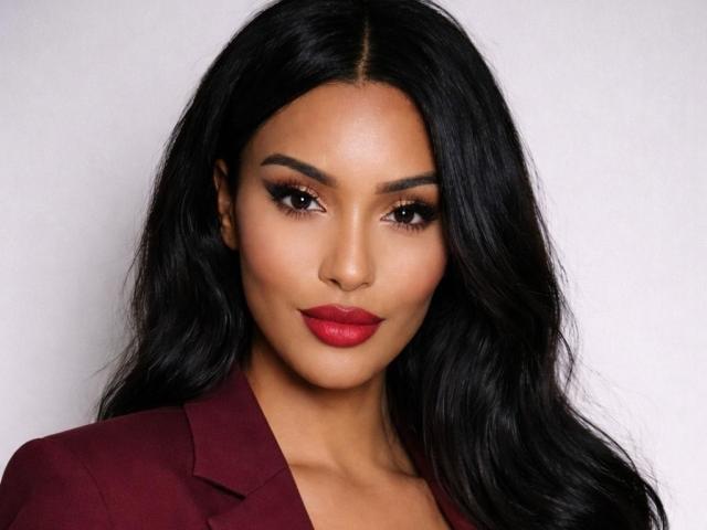 Aleciax has long, wavy black hair. Her face is centered, with a serious expression and dark, almond-shaped eyes. She has bold, arched eyebrows and wears dark red lipstick. Aleciax's skin is smooth and has a warm, brownish tone. She wears a maroon blazer with a slightly open collar. The lighting is even, highlighting her facial features. The image is a close-up portrait. Her makeup is dramatic, with dark eyeliner and well-defined eyebrows. Her hair is glossy and flows to the sides.