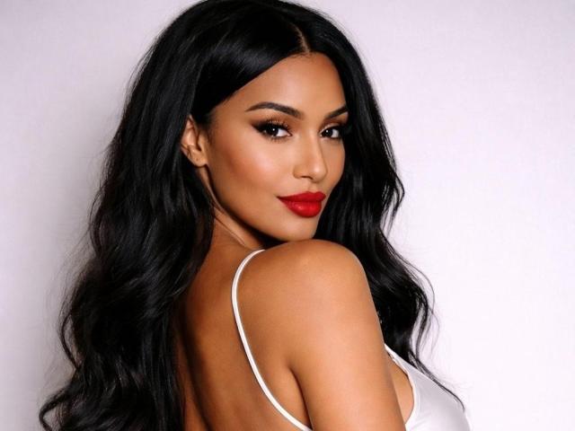 Aleciax has long, wavy black hair and a medium skin tone. She looks over her shoulder with a slight smile, wearing bright red lipstick. Her dark, defined eyebrows contrast with her smooth, even skin. She's dressed in a white spaghetti-strap top, showing her bare shoulders. The composition focuses on her upper body and face, with soft lighting highlighting her facial features. The image is sharp with a slight shadow on the right, emphasizing her hair's texture.