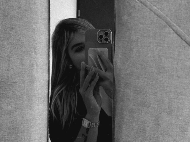 Black and white selfie of SashaPolet. SashaPolet, with long, wavy, light brown hair, holds a smartphone in front of her face, partially obscuring her features. She has dark, arched eyebrows and wears a watch on her left wrist. Her right hand, with manicured nails, holds the phone. The phone has a triple-camera setup on the back. The image is framed by two vertical, textured fabric panels. The overall composition is tight, focusing on SashaPolet's face and hand. The texture of the fabrics contrasts with the smooth phone and SashaPolet's hair.