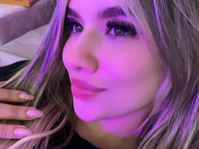 Close-up portrait of SashaPolet with purple-tinged lighting. SashaPolet has long, straight, blonde hair with darker roots, and is looking at the camera with a slight, confident smile. She has dark, thick eyebrows, long, curled eyelashes, and purple eye shadow. Her lips are full and painted pink. SashaPolet's right hand, with light pink nail polish, rests near her face. The image has a soft, purple glow, highlighting her facial features. The background is blurred, focusing attention on SashaPolet's face and expression.