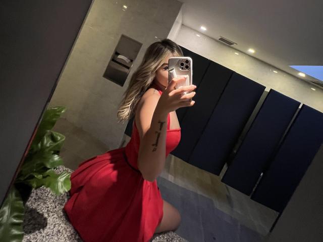 SashaPolet takes a selfie in a mirror. She's kneeling on a carpeted floor, wearing a bright red dress with a flared skirt that accentuates her curves. She holds a silver iPhone with a pink case in her right hand, partially covering her face. She has blonde hair with dark roots and a tattoo of three vertical lines on her left forearm. The background includes a plant and dark blue stall dividers. The image is well-lit with warm lighting.